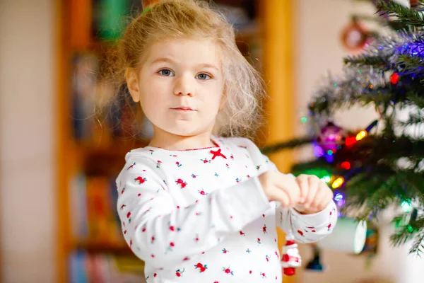 Imádnivaló kislány pizsamában karácsonyfát díszít játékkal a cuki kezekben. Egy kisgyerek hálóingben a karácsonyfa mellett. ünneplése hagyományos családi téli nyaralás — Stock Fotó