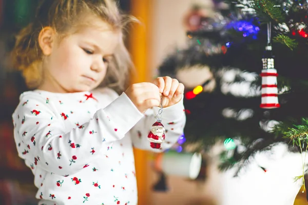Roztomilé batole dívka v pyžamu zdobí vánoční stromek s hračkou v roztomilých rukou. Malé dítě v noční košili stojící u vánočního stromku. oslava tradiční rodinné zimní dovolené — Stock fotografie