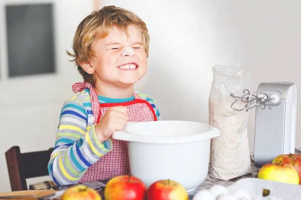 Bonito pequeno menino pré-escolar loiro feliz cozinhando bolo de maçã e muffins na cozinha doméstica. Engraçado adorável criança saudável se divertindo com o trabalho com misturador, farinha, ovos, frutas. Pequeno ajudante dentro de casa — Fotografia de Stock