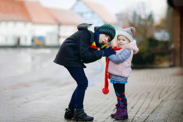 School kid boy and brother warming hands of little sister, toddler girl on cold snowy grey winter day. Family, two lovely siblings children playing with snow, outdoors during snowfall. — Stock Photo, Image