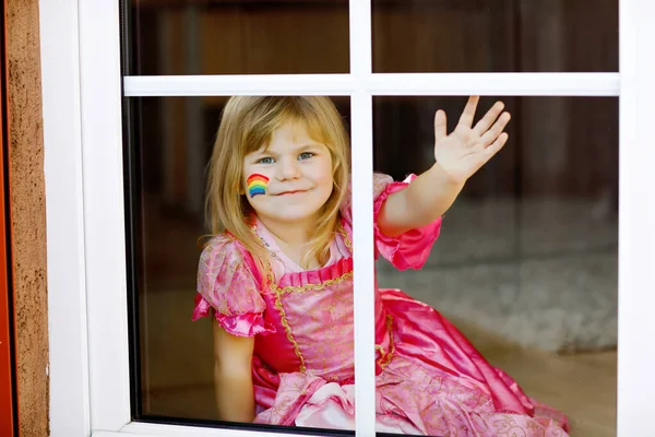 Niedliche einsame kleine Mädchen in Prinzessinnenkleid am Fenster sitzend mit Regenbogen mit bunten Farben im Gesicht während der Coronavirus-Quarantäne. Kinder basteln und bemalen Regenbogen auf der ganzen Welt — Stockfoto