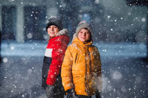 Zwei kleine Jungen der Grundschulklasse laufen bei Schneefall am frühen Morgen zur Schule. Glückliche Kinder, die Spaß haben und mit dem ersten Schnee spielen. Geschwister und Freunde in bunter Winterkleidung — Stockfoto