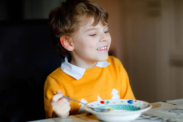 小学生の男の子が野菜スープを屋内で食べる。家庭用キッチンや学校の食堂で金髪の子供。かわいい子供と健康食品、麺と有機ビーガンスープ、鶏や野菜. — ストック写真