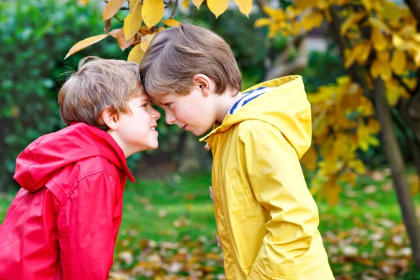 Dos pequeños mejores amigos y niños parque de otoño en ropa de colores. Felices hermanos niños divirtiéndose en abrigos de lluvia rojos y amarillos y botas de goma. Familia jugando al aire libre. ocio activo. — Foto de Stock