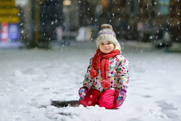 En söt liten flicka som går utomhus på vintern. Söta småbarn under starkt snöfall på kvällen. Barn som har kul med snö. Bär varm baby färgglada kläder och hatt med kulor. — Stockfoto