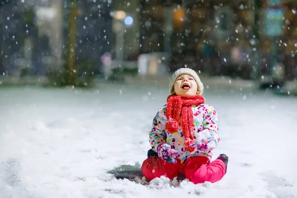 冬に屋外を歩く愛らしい小さな幼児の女の子。夕方の強い降雪の間にかわいい幼児。雪と遊ぶ子供。暖かい赤ちゃんのカラフルな服やボブル付きの帽子を身に着けて. — ストック写真