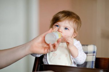 Şişeden mama içen tatlı bir kız bebek. Yemek, çocuk, beslenme ve insanların konsepti - tatlı bebek, kaşıkla sandalyede oturan ve evde yemek yiyen kız.