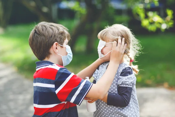 El chico de la escuela ayudando a la hermanita a poner máscara médica. Chica sosteniendo spray higiénico. Hermano y linda niña aprenden reglas de higiene. Familia durante el bloqueo del coronavirus. Protección de los ávidos. — Foto de Stock