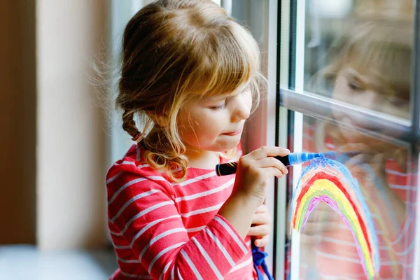 Adoralbe little toddler girl with rainbow painted with colorful window color during pandemic coronavirus quarantine. Child painting rainbows and hearts around the world with words Lets all be well. — Stock Photo, Image