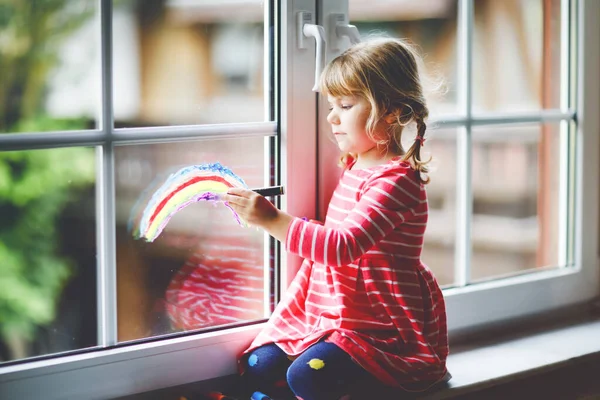 Adoralbe kleine peuter meisje met regenboog geschilderd met kleurrijke venster kleur tijdens pandemische coronavirus quarantaine. Kind schilderen regenbogen en harten over de hele wereld met woorden Laten we allemaal goed zijn. — Stockfoto