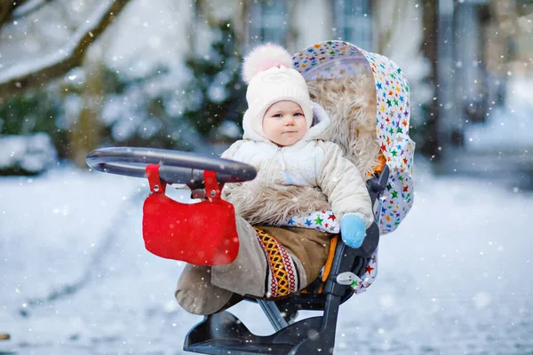 Söt liten vacker liten flicka sitter i barnvagn eller barnvagn på kall snöig vinterdag. Glad leende barn i varma kläder, mode snygg baby coat. Babys första snö. — Stockfoto