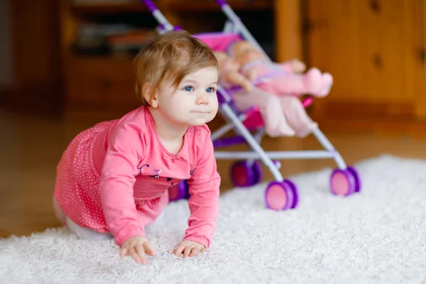 Schattig schattig baby meisje kruipen en spelen met pop rijtuig. Prachtig peuterkind met kinderwagen met kinderspeeltje thuis. Gelukkig kind met twee poppen — Stockfoto