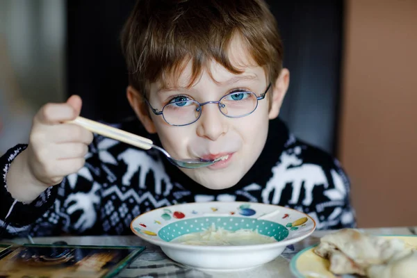 Kleiner Schuljunge mit Brille, der drinnen Gemüsesuppe isst. Blondes Kind in der heimischen Küche oder in der Schulkantine. Nettes Kind und gesunde Kost, Bio-vegane Suppe mit Nudeln, Huhn und Gemüse. — Stockfoto