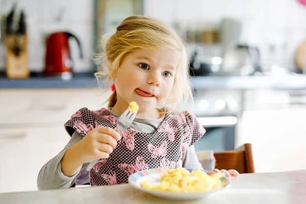 Schöne Kleinkind Mädchen essen gesunde Bratkartoffeln zum Mittagessen. Nettes glückliches Baby in bunten Kleidern, das in der Küche des Hauses, der Kita oder des Kinderzimmers sitzt. Kind isst Gemüse. — Stockfoto