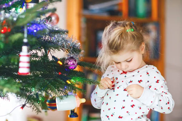 Urocza dziewczynka maluch w piżamie dekorowanie choinki z zabawką w uroczych rękach. Małe dziecko w koszuli nocnej stojące przy choince świątecznej. obchody tradycyjnego rodzinnego zimowego urlopu — Zdjęcie stockowe