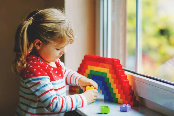 Linda niña pequeña por ventana crear arco iris con bloques de plástico de colores durante la cuarentena pandémica coronavirus. Los niños hicieron y pintaron arcoíris alrededor del mundo como signo. — Foto de Stock