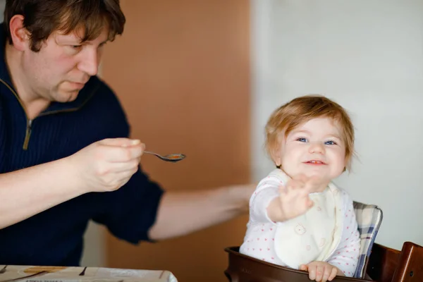 Отец кормит девочку из ложечного пюре и овощей. food, child, feeding and people concept-cute toddler, daughter and dad with spoon sitting in highchair and eating at home. — стоковое фото