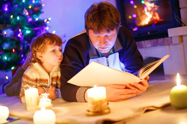 Vater und süßer kleiner Junge lesen Buch am Kamin, Kerzen und Kamin. — Stockfoto