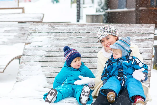 Jonge moeder met twee kleine peuterjongens zittend op de bank in het winterpark. Vrouw spelen en knuffelen met schattige baby zonen. — Stockfoto