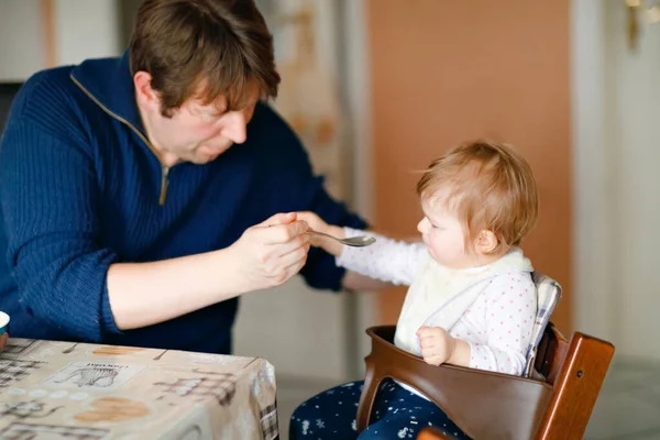 Отец кормит девочку из ложечного пюре и овощей. food, child, feeding and people concept-cute toddler, daughter and dad with spoon sitting in highchair and eating at home. — стоковое фото