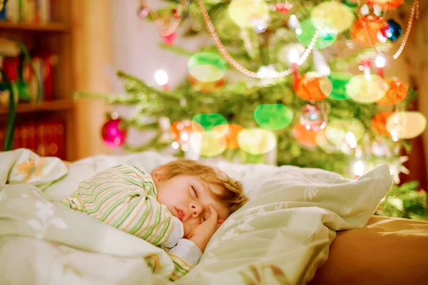Little cute blond boy sleeping under Christmas tree and dreaming of Santa at home, indoors. Traditional Christian festival. Happy kid child waiting for gifts on xmas. Cozy soft light — Stock Photo, Image