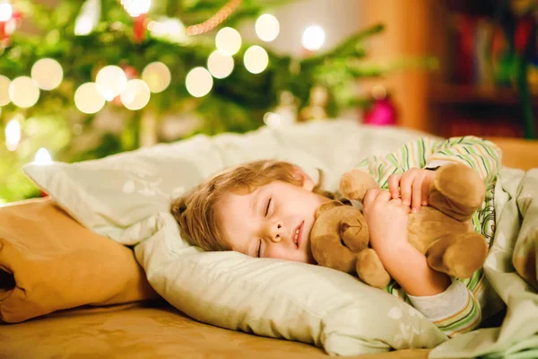 Little cute blond boy sleeping under Christmas tree and dreaming of Santa at home, indoors. Traditional Christian festival. Happy kid child waiting for gifts on xmas. Cozy soft light — Stock Photo, Image