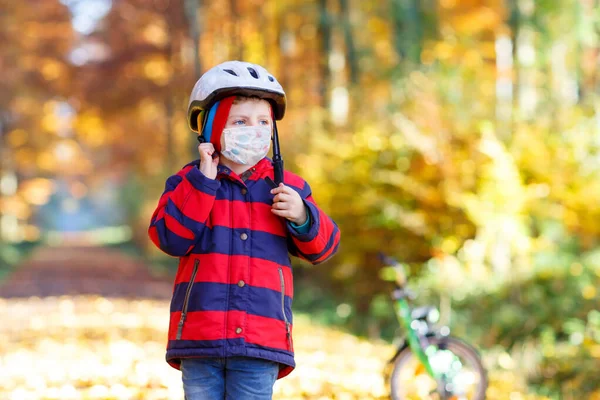 医療用マスクを着た小さな男の子が自転車用の安全ヘルメットを頭の上に置きます。秋の森公園で自転車で。安全性、スポーツ、子供の概念とレジャーコロナウイルスのロックダウン中に. — ストック写真