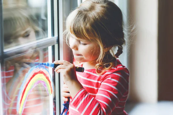 Adoralbe little toddler girl with rainbow painted with colorful window color during pandemic coronavirus quarantine. Child painting rainbows and hearts around the world with words Lets all be well. — Stock Photo, Image