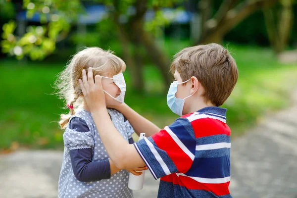 El chico de la escuela ayudando a la hermanita a poner máscara médica. Chica sosteniendo spray higiénico. Hermano y linda niña aprenden reglas de higiene. Familia durante el bloqueo del coronavirus. Protección de los ávidos. — Foto de Stock