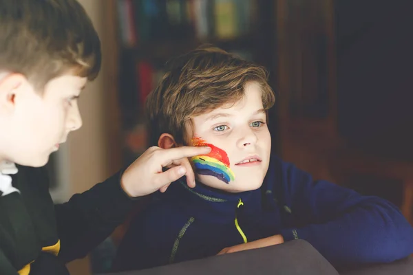 Twee jongens met regenboog met kleurrijke kleuren op het gezicht tijdens pandemische coronavirus quarantaine. kind schildert het gezicht van broer. Kinderen maken regenbogen over de hele wereld. Broers en zussen, beste vrienden — Stockfoto