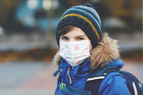 Niño pequeño con máscara médica camino a la escuela. Mochila para niños. Colegial en otoño frío o día de invierno con ropa de abrigo. Tiempo de bloqueo y cuarentena durante la enfermedad pandémica de corona — Foto de Stock