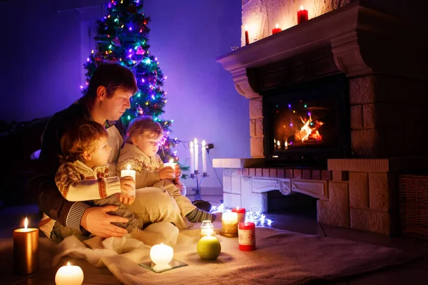 Far och två små småpojkar sitter vid skorstenen, ljus och öppen spis och tittar på eld. Familjen firar jul. Med julgran och ljus på bakgrunden. Barn glada över gåvor — Stockfoto