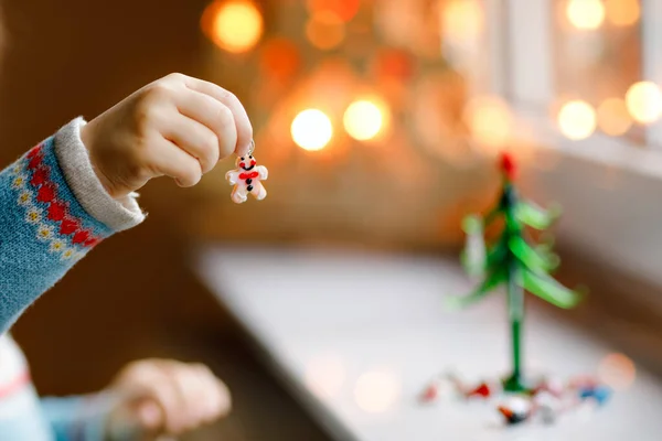 Primer plano de niña por ventana y decorando pequeño árbol de Navidad de vidrio con pequeños juguetes de Navidad. El niño celebra la fiesta familiar tradicional. Primer plano en las manos con el hombre de jengibre — Foto de Stock