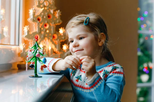 Kleine Mädchen sitzen am Fenster und schmücken kleine gläserne Weihnachtsbäume mit winzigem Weihnachtsspielzeug. Glückliches gesundes Kind feiert traditionellen Familienurlaub. Liebenswertes Baby. — Stockfoto