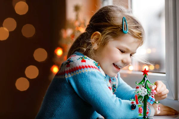 Niña pequeña sentada junto a la ventana y decorando un pequeño árbol de Navidad de vidrio con pequeños juguetes de Navidad. Feliz niño sano celebrar la fiesta tradicional familiar. Adorable bebé.. — Foto de Stock