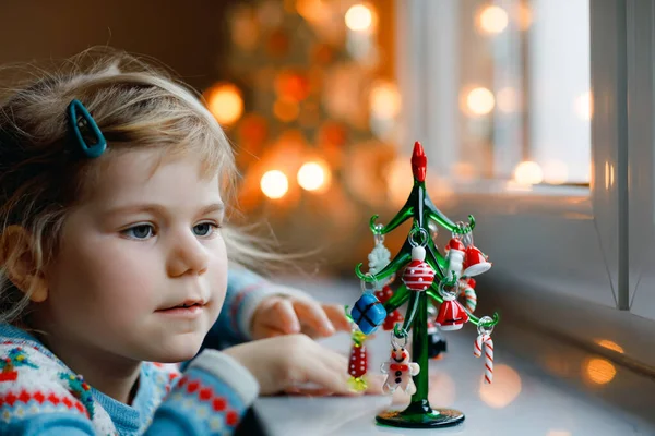 Kleine Mädchen sitzen am Fenster und schmücken kleine gläserne Weihnachtsbäume mit winzigem Weihnachtsspielzeug. Glückliches gesundes Kind feiert traditionellen Familienurlaub. Liebenswertes Baby. — Stockfoto