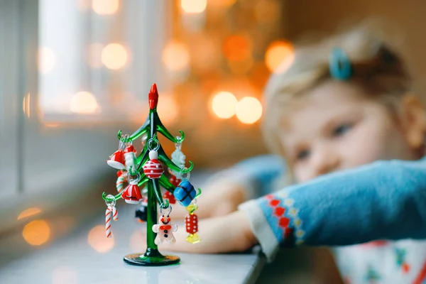 Dziewczynka Litte Maluch siedzi przy oknie i dekoracji małej choinki szkła z małych zabawek xmas. Szczęśliwe zdrowe dziecko świętować rodzinne tradycyjne wakacje. Urocze dziecko. Selektywna koncentracja na drzewie — Zdjęcie stockowe