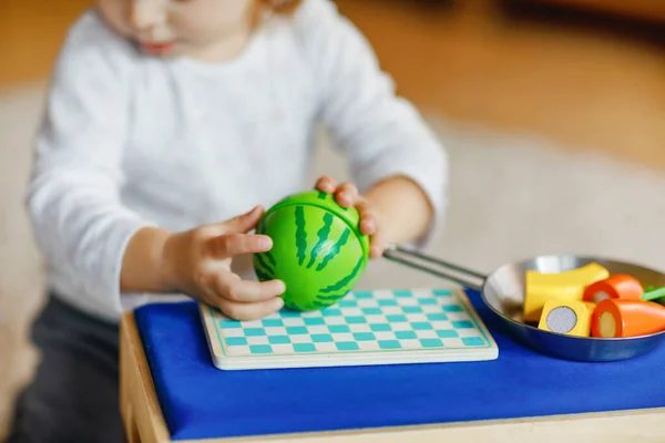 Nettes kleines Mädchen, das zu Hause mit Öko-Holzspielzeug spielt. Glückliches gesundes aufgeregtes Kind, das Gemüse und Obst mit dem Spielzeugmesser schneidet. Baby-Mädchen haben Spaß mit Rolle Küche und Kochen Spiel. — Stockfoto