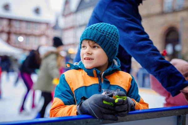 Glad liten pojke i färgglada varma kläder på skridskobana av julmarknad eller rättvis dricka varm punch eller choklad. Friska barn som har roligt på skridskor. personer som har aktiv vinterfritid — Stockfoto