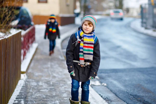 雪の中、小学生の男の子2人が学校に歩いています。楽しい時間を過ごし、初めての雪で遊ぶ幸せな子供たち。兄弟姉妹友人でバックパックでカラフルな冬服. — ストック写真