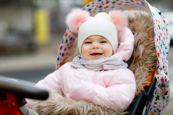 Schattig klein mooi babymeisje zittend in de kinderwagen of kinderwagen op koude herfst-, winter- of lentedag. Gelukkig lachend kind in warme kleren, stijlvolle babyjas en hoed. Sneeuw valt neer — Stockfoto