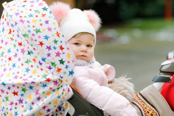 Cute little beautiful baby girl sitting in the pram or stroller on cold autumn, winter or spring day. Happy smiling child in warm clothes, fashion stylish baby coat and hat. Snow falling down — Stock Photo, Image