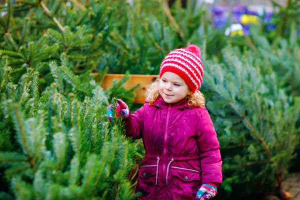 Adorable niñita sosteniendo el árbol de Navidad en el mercado. Niño feliz bebé sano en ropa de moda de invierno elegir y comprar gran árbol de Navidad en la tienda al aire libre. Familia, tradición, celebración. —  Fotos de Stock