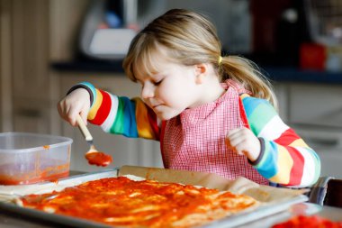 Sevimli küçük kız evde İtalyan pizzası yapıyor. Ev mutfağında, evde eğlenen sevimli mutlu çocuk. Çocuk, anaokulu öğrencisi sağlıklı yemek hazırlıyor ve yardım ediyor.