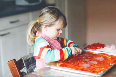 Sevimli küçük kız evde İtalyan pizzası yapıyor. Ev mutfağında, evde eğlenen sevimli mutlu çocuk. Çocuk, anaokulu öğrencisi sağlıklı yemek hazırlıyor ve yardım ediyor.