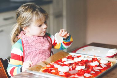 Sevimli küçük kız evde İtalyan pizzası yapıyor. Ev mutfağında, evde eğlenen sevimli mutlu çocuk. Çocuk, anaokulu öğrencisi sağlıklı yemek hazırlıyor ve yardım ediyor.