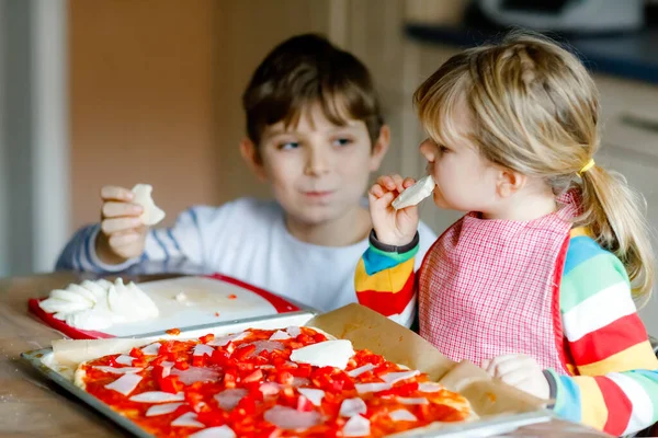 2人の兄弟、小さな子供たちが自宅でイタリアのピザを作っています。かわいい幼児の女の子と学校の少年は、屋内で自宅のキッチンで楽しんでいます。兄弟姉妹、家族は食事の手伝いや準備をする — ストック写真