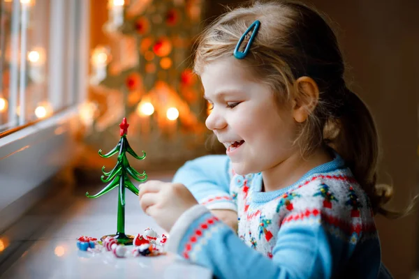 Litte småbarn flicka sitter vid fönstret och dekorera små glas julgran med små xmas leksaker. Glad frisk barn fira familjens traditionella semester. Förtjusande barn. — Stockfoto