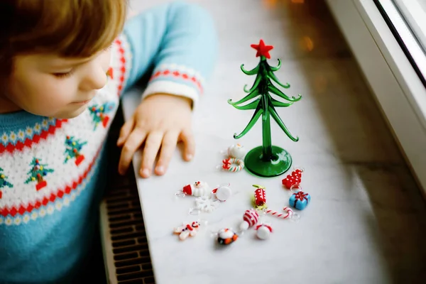 Closeup of toddler girl by window and decorating small glass Christmas tree with tiny xmas toys. Child celebrate family traditional holiday. Close-up on hands with gingerbread man — Stock Photo, Image