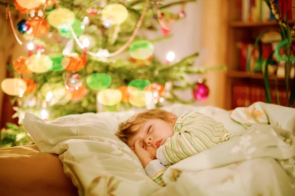 Little cute blond boy sleeping under Christmas tree and dreaming of Santa at home, indoors. Traditional Christian festival. Happy kid child waiting for gifts on xmas. Cozy soft light — Stock Photo, Image